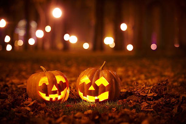 Two jack-o '- lanterns sitting in the grass with lights behind them.