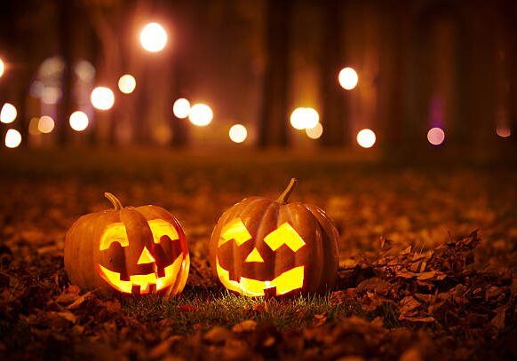 Two jack-o '- lanterns sitting in the grass with lights behind them.